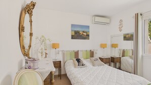 A bedroom with a large bed, two bedside tables, a chair, a mirror, a vase with flowers, a picture on the wall, and an air conditioning unit.