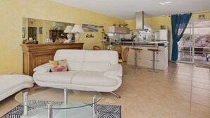 A living room with a white sofa, a glass coffee table, and a kitchen area with a stove and cabinets.