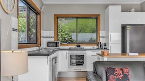 A compact kitchen with a microwave, stove, and oven, a wooden window, and a black leather sofa with a cushion. - Hillcrest Retreat with River Views (Tweed Heads South)