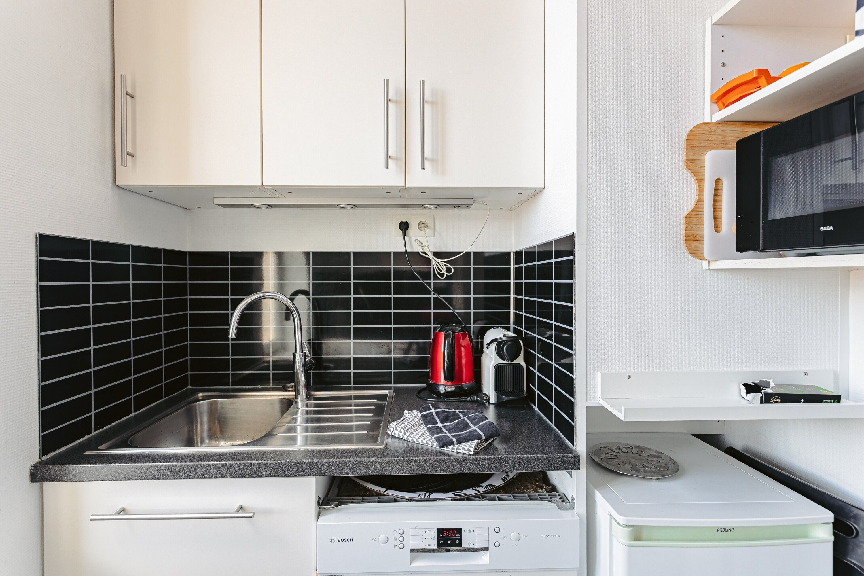 Une cuisine compacte avec des meubles blancs, un dosseret de carrelage noir, un évier en acier inoxydable, un lave-vaisselle Bosch et un micro-ondes posé sur une étagère en bois.