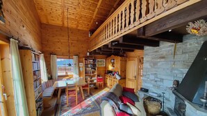 Interior de una cabaña de madera con comedor, estanterías, un sofá con cojines, chimenea y pared de piedra.