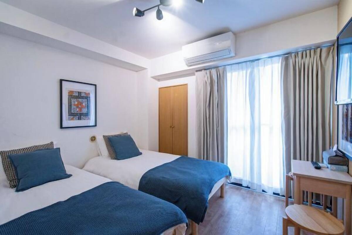 A hotel room with two beds, a desk, an air conditioning unit, a door, and a window with curtains.