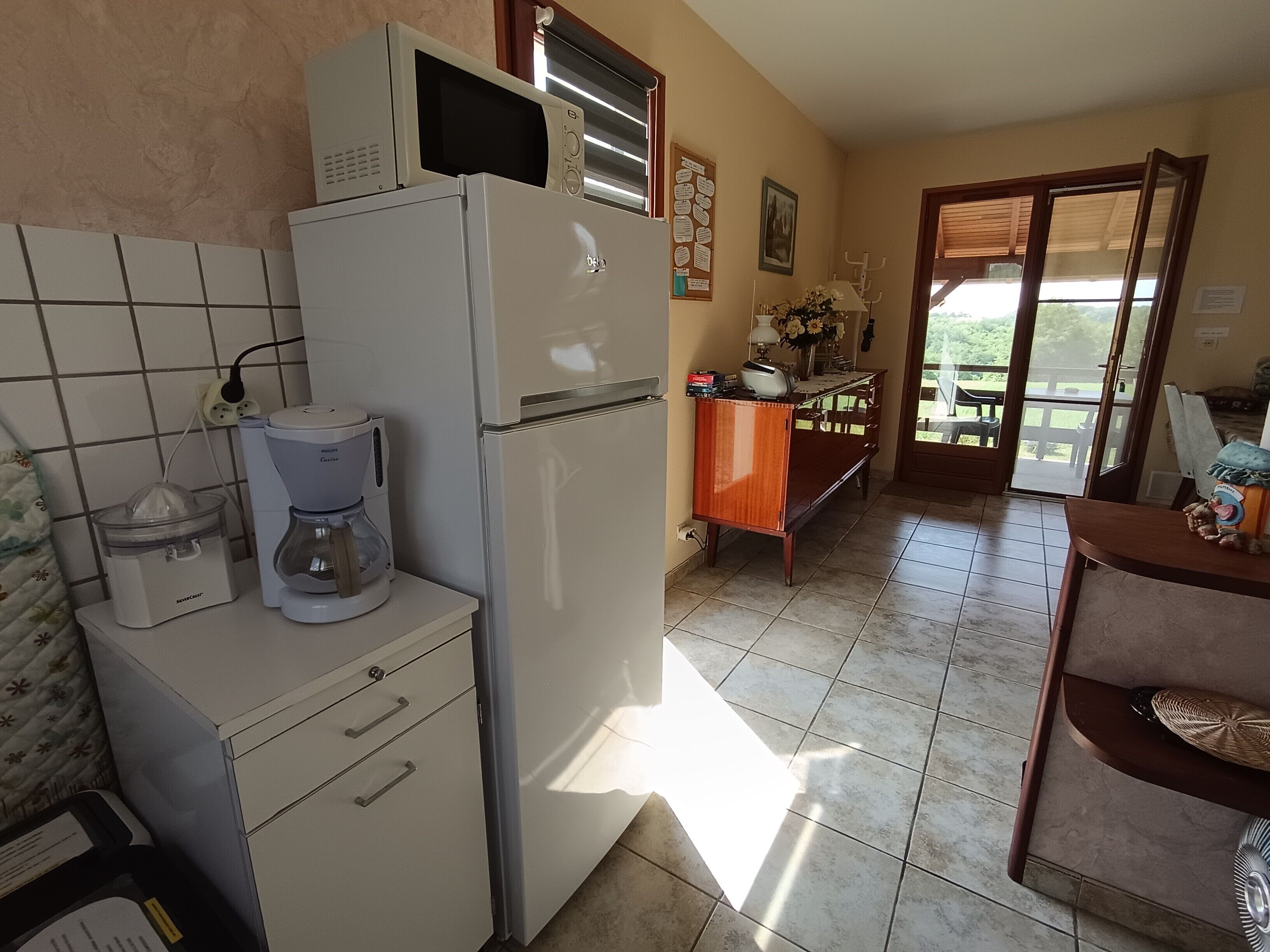 Une cuisine équipée d’un réfrigérateur, d’une machine à café et d’un four à micro-ondes. On y trouve un meuble en bois orné d’un vase et de fleurs, et une porte vitrée offre une vue sur l’extérieur.
