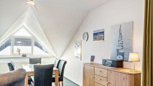A room with a dining table, chairs, a sofa, a wooden cabinet, a radio, a clock, and a framed picture on the wall. - Apartment 'Kirschner's 2' with Shared Terrace and Wi-Fi (Horumersiel-Schillig)