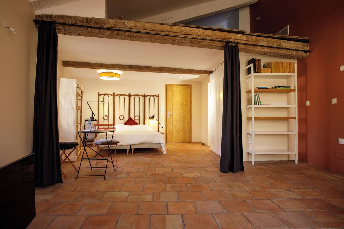 A bedroom with a bed, a chair, a table, a wooden door, and a bookshelf.