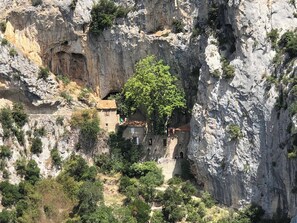 A small building nestled in a rocky cliffside with a large tree in front. - Ostal de la placette (Padern)