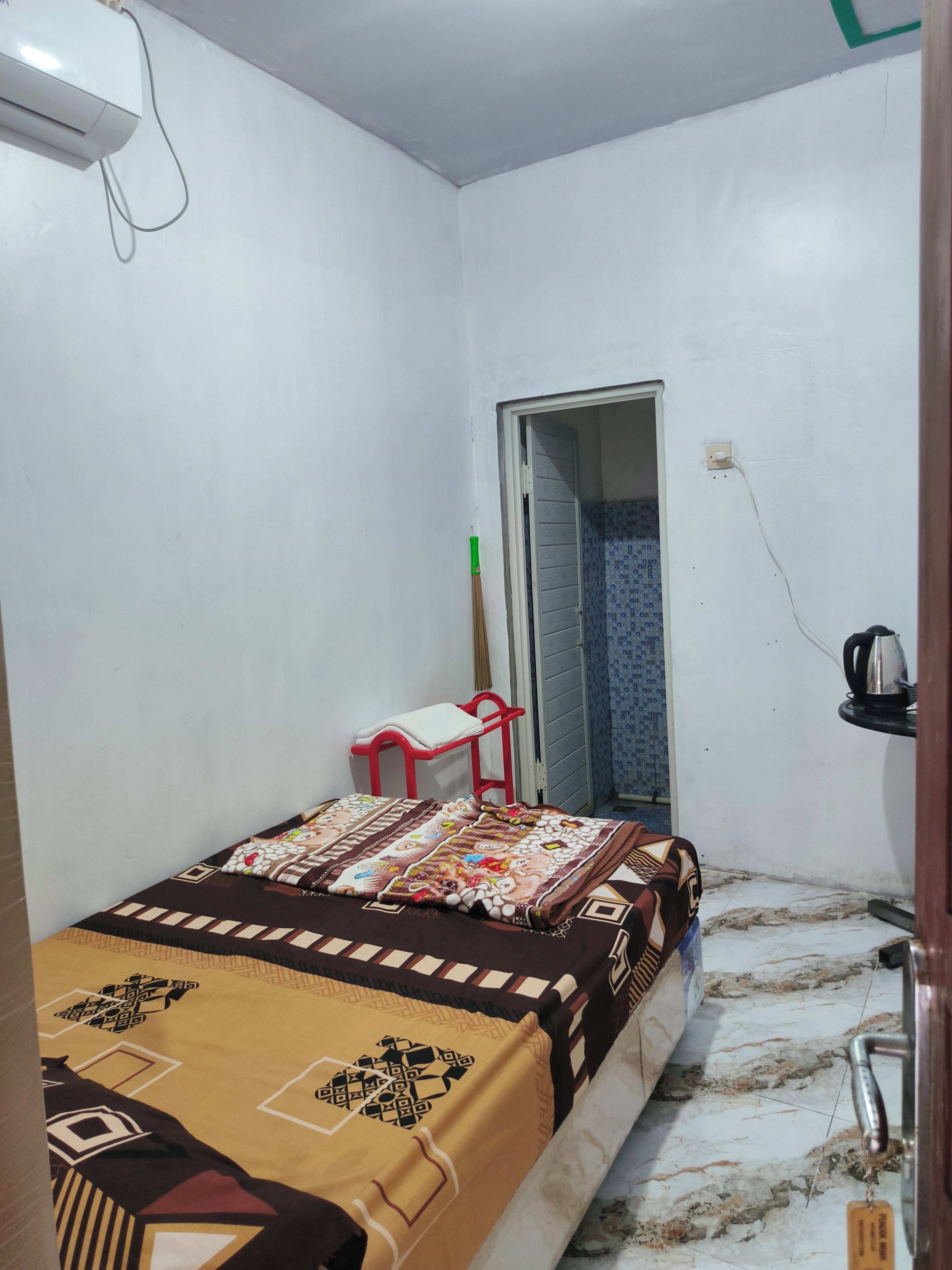 A small, sparsely furnished room with a bed, a red chair, a white air conditioner, and a door leading to a bathroom.