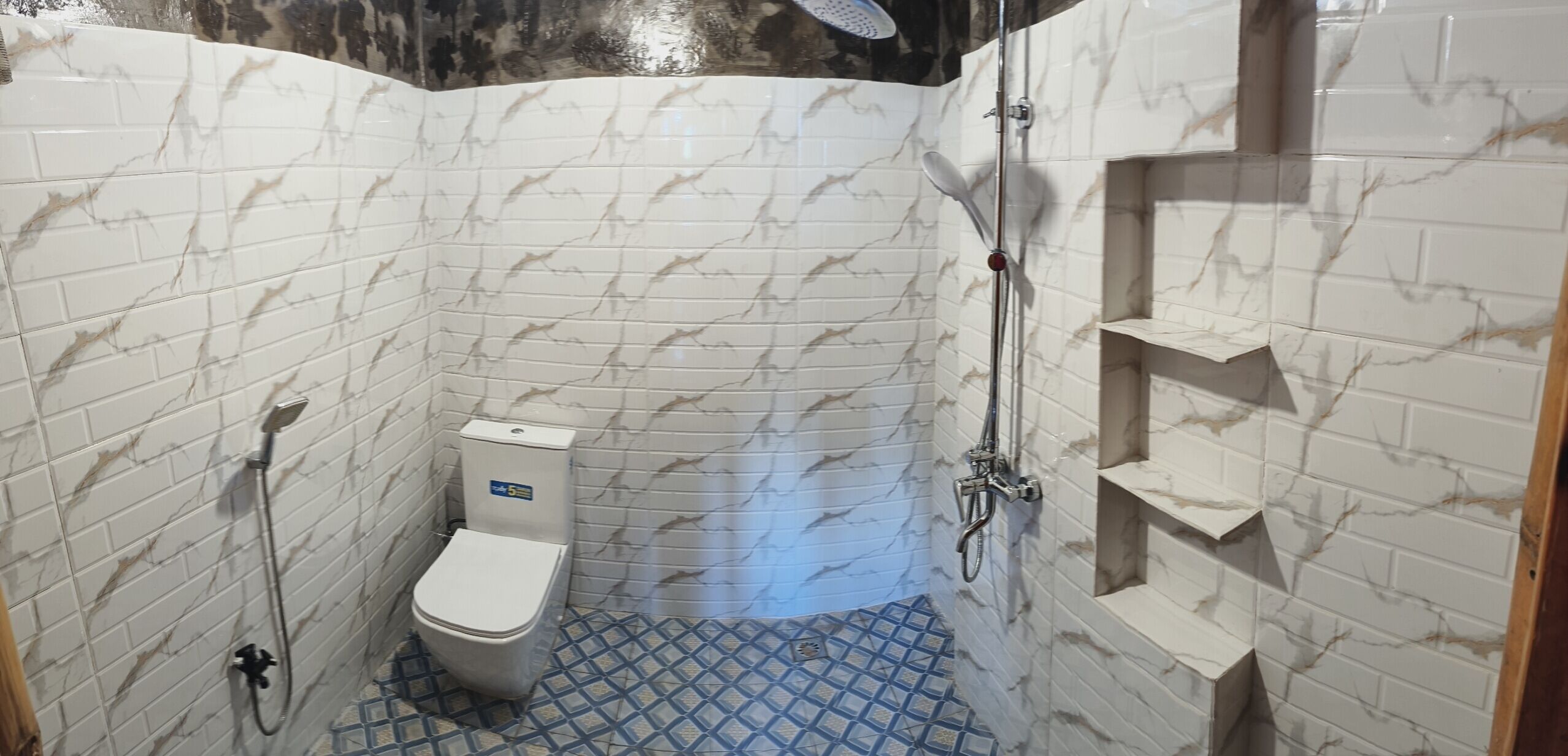 A small, tiled bathroom with a toilet, shower, and a shelf unit.