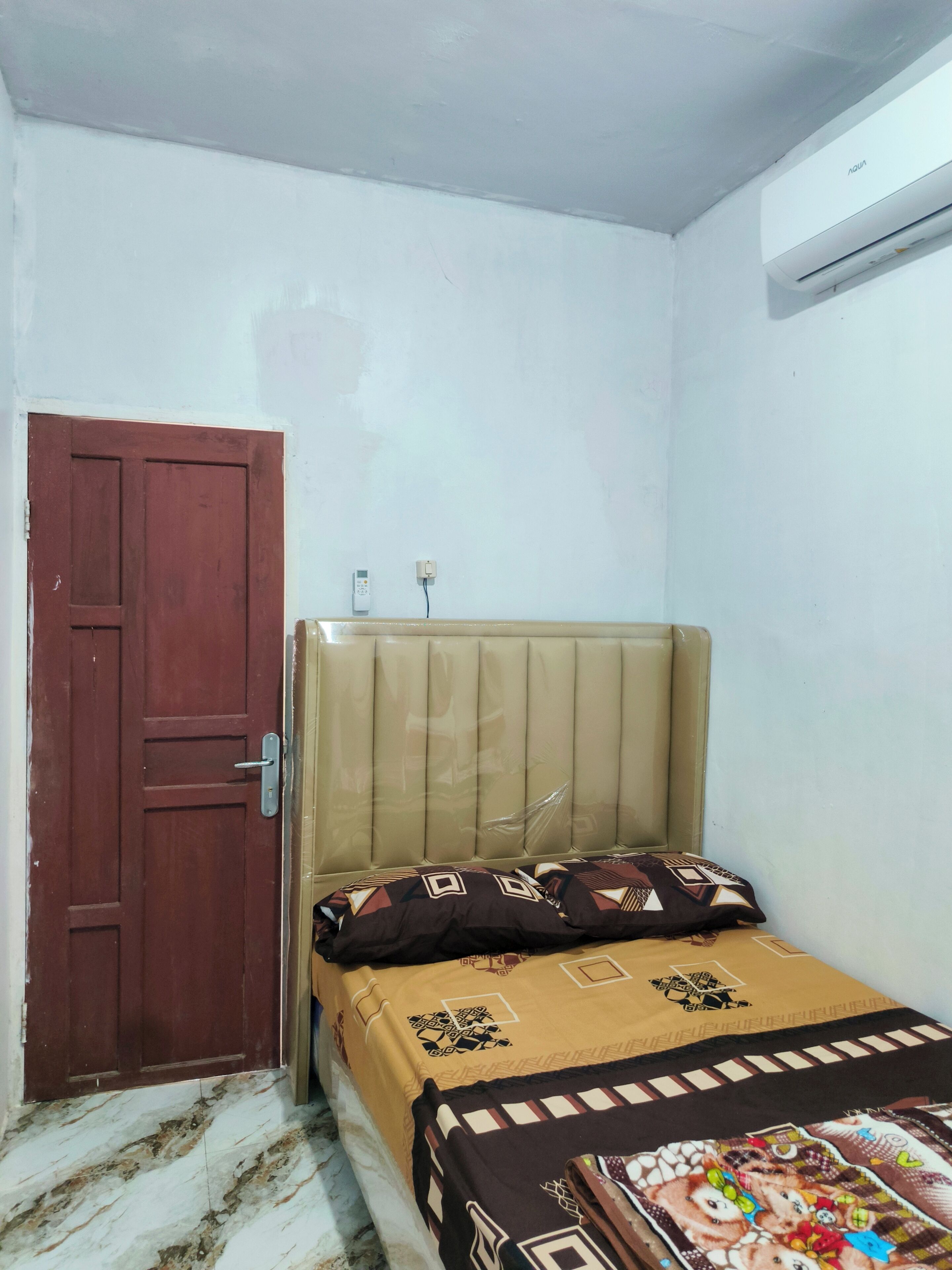 A bedroom with a wooden door, a bed with patterned bedding, a wall-mounted air conditioner, and a marble floor.