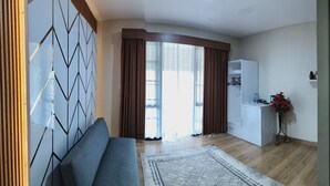 A modern living room with a sofa, a patterned rug, a wooden wall panel, a kitchen area with a white cabinet, and a potted plant. - Pondok Indah At Teluk Asmara Beach (Sitiardjo)