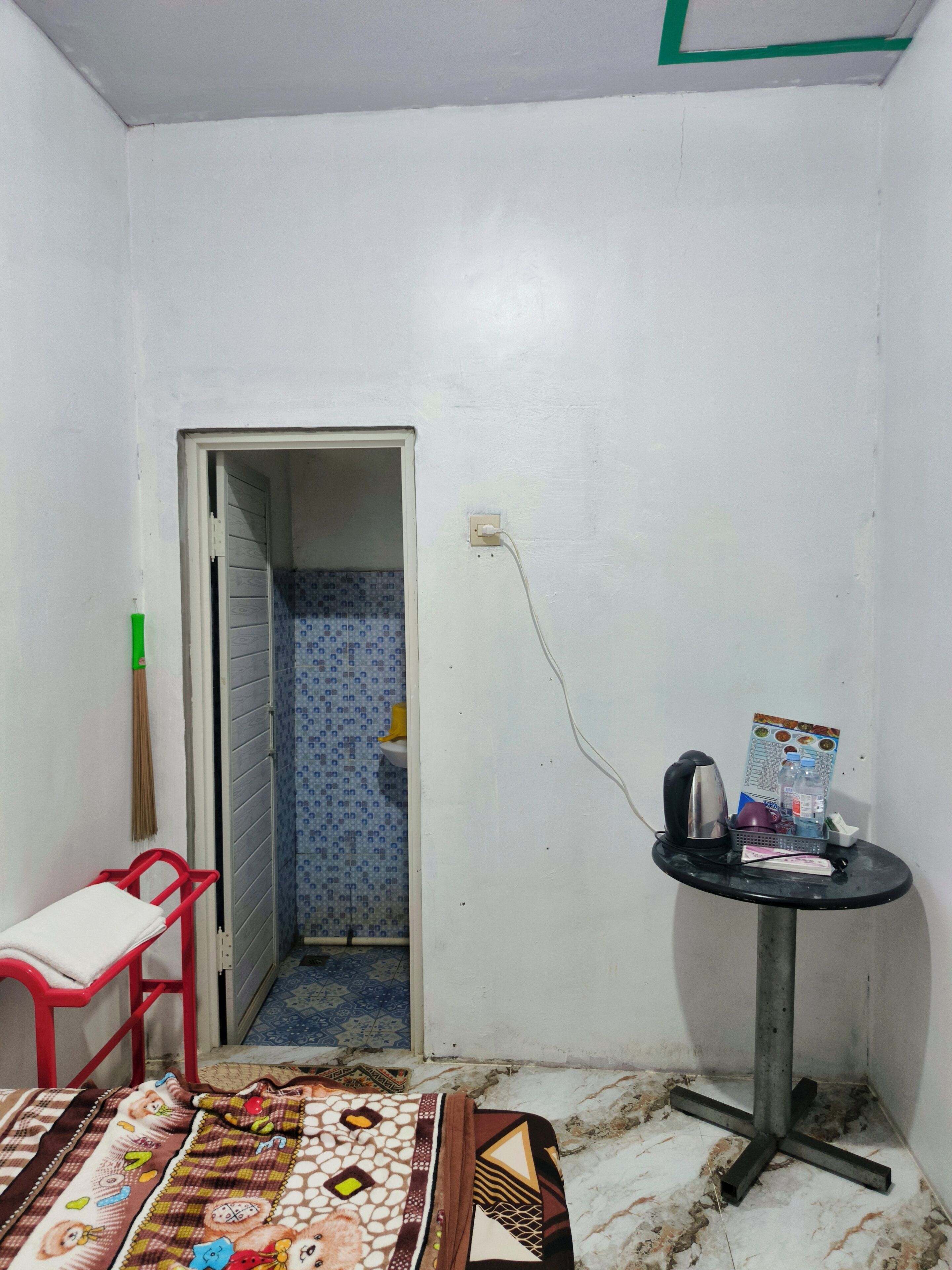 Une petite pièce étroite, avec un lit simple, une chaise rouge, une petite table sur laquelle se trouvent une bouilloire et une bouteille d’eau, et une porte donnant sur une salle de bain dont le sol est recouvert d’un motif bleu.
