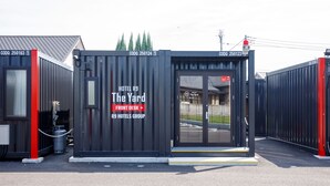 Entrada moderna de un hotel con puerta de cristal, un letrero que dice 'The Yard' y 'R9 Hotels Group', y un pilar rojo.