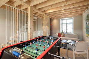 Een kamer met een tafelvoetbaltafel, een bank, een raam met jaloezieën en een schilderij aan de muur.