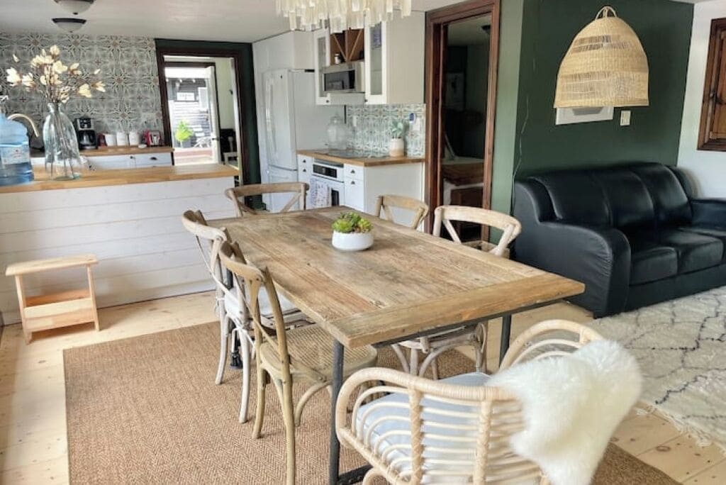 Ein Essbereich mit Holztisch und Korbstühlen, eine schwarze Ledersofa und eine Küche im Hintergrund.