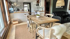 A dining area with a wooden table and wicker chairs, a kitchen with white cabinets, and a black sofa in the living area. - Muskoka Cabana - Sandy beach cottage in Muskoka (Utterson)