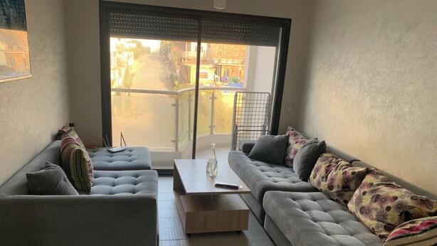 A modern living room with a grey sofa set, a wooden coffee table, and a large sliding glass door leading to an outdoor area. - Nice apartment (Kenitra)