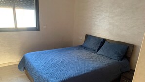A single bed with a blue quilt in a room with a plain wall and a window. - Nice apartment (Kenitra)