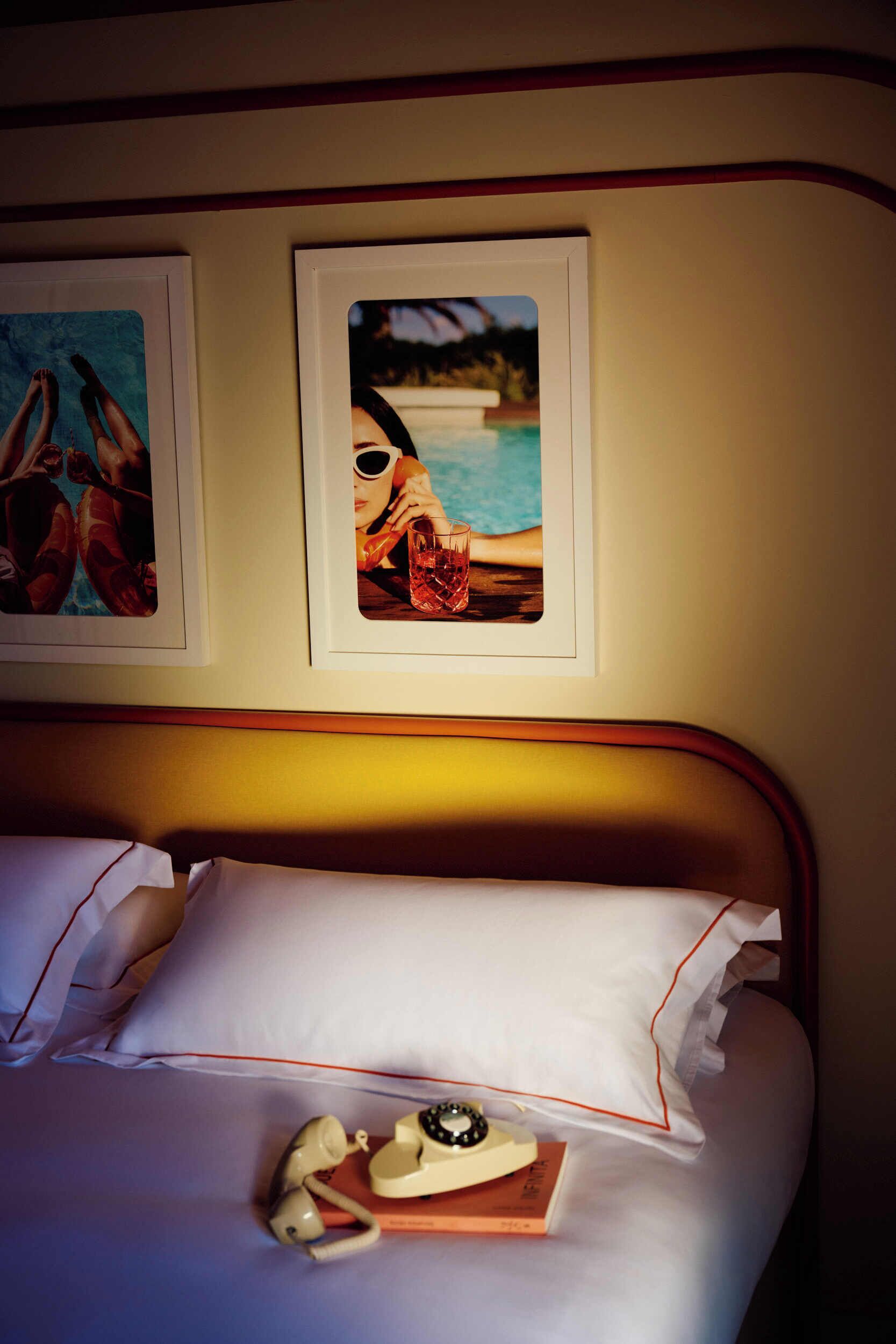 Una cama bien hecha con sábanas blancas y un cabecero de madera, un cuadro enmarcado de un paisaje tropical, y una mesita de noche con un teléfono y un libro.