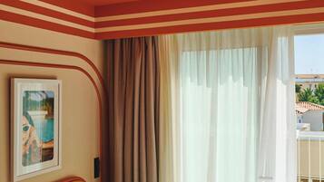 Quarto de hotel com cama, cortinas, um quadro emoldurado e uma varanda com guarda-corpo.