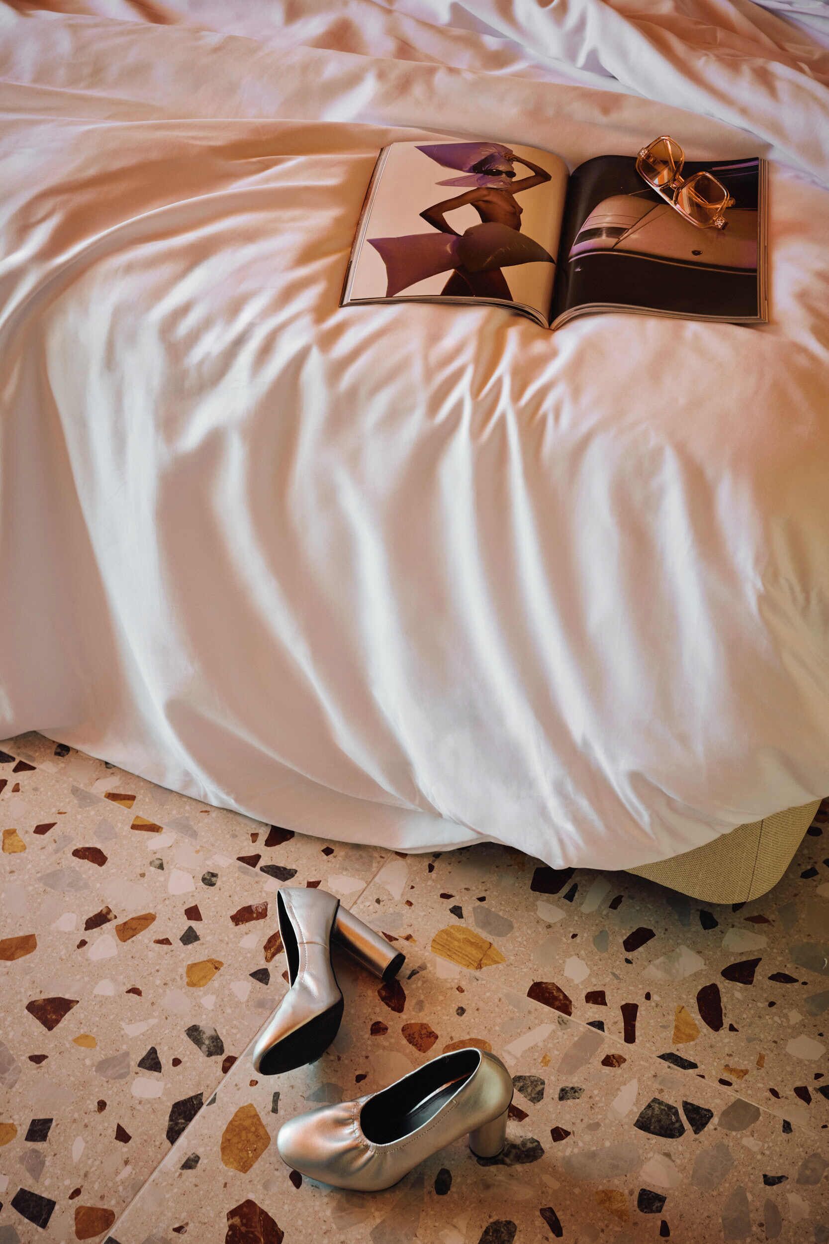 Una cama con una colcha blanca, un libro y un par de zapatos de tacón plateados sobre un piso de terrazo.