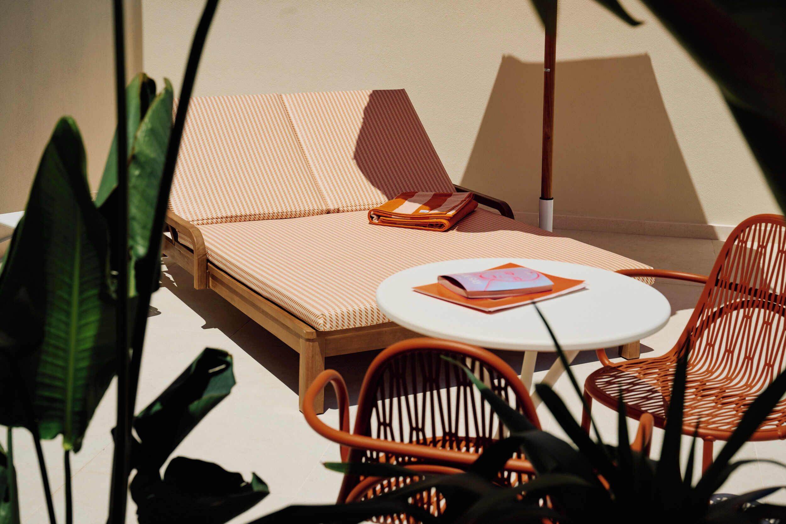 A sun lounge with a cushioned bench, a small round table with a book, and wicker chairs.