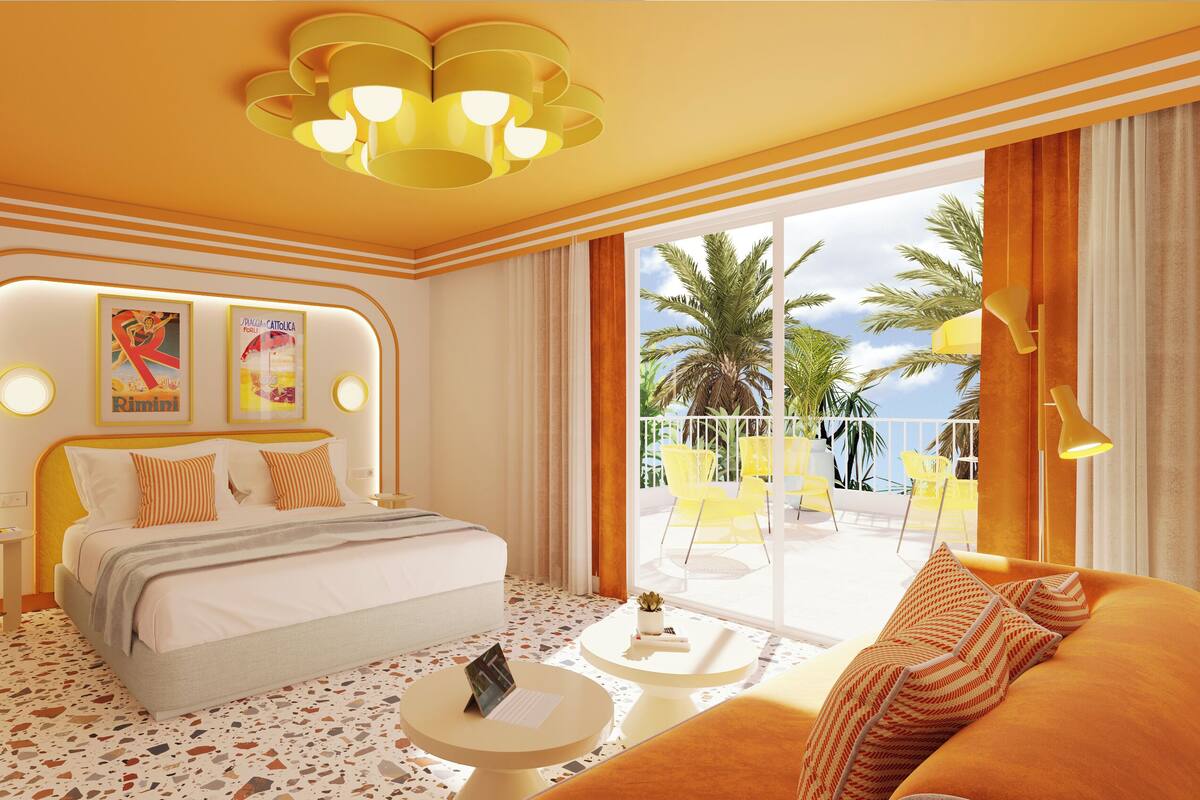 A hotel room with a bed, a sofa, a small table, a balcony with yellow chairs, and a view of palm trees.