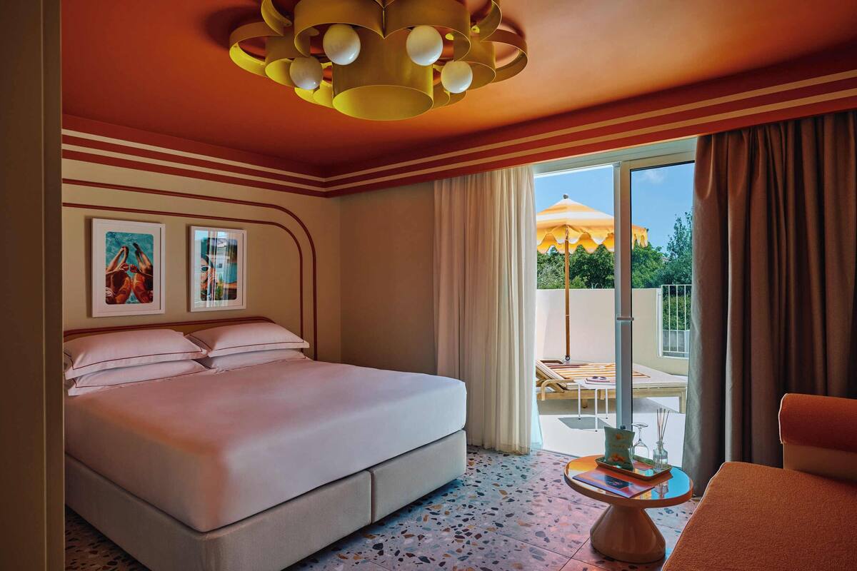 A hotel room with a bed, a small table, a chair, a ceiling light fixture, and a view of the outdoors.