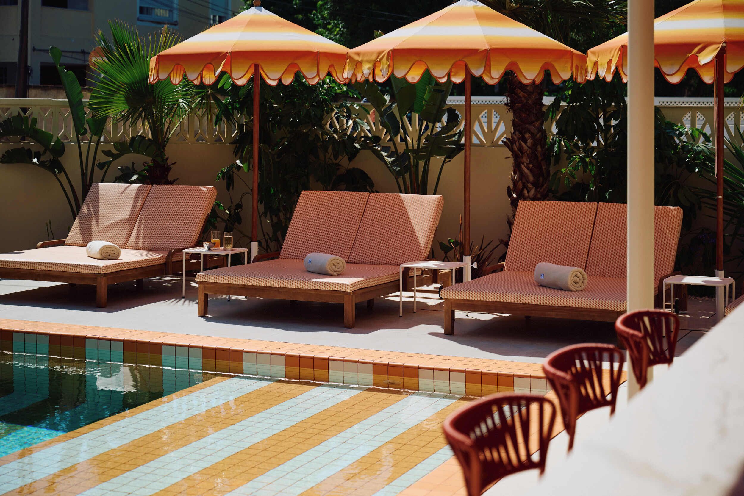 Zona junto a la piscina con sillas de descanso y sombrillas, una piscina con baldosas y vegetación circundante.