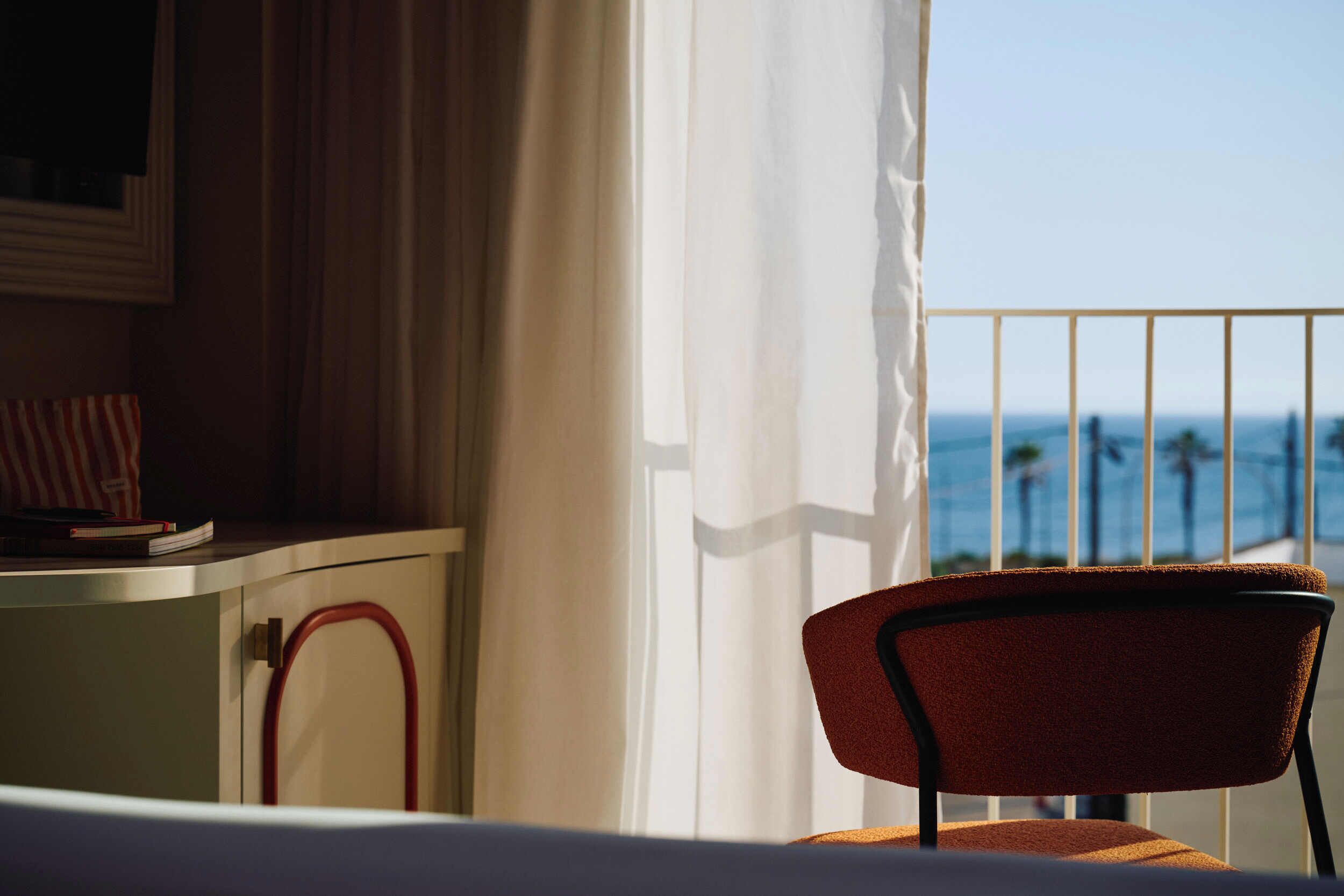 Une pièce avec vue sur l’océan, une chaise, un bureau avec un livre et un rideau.