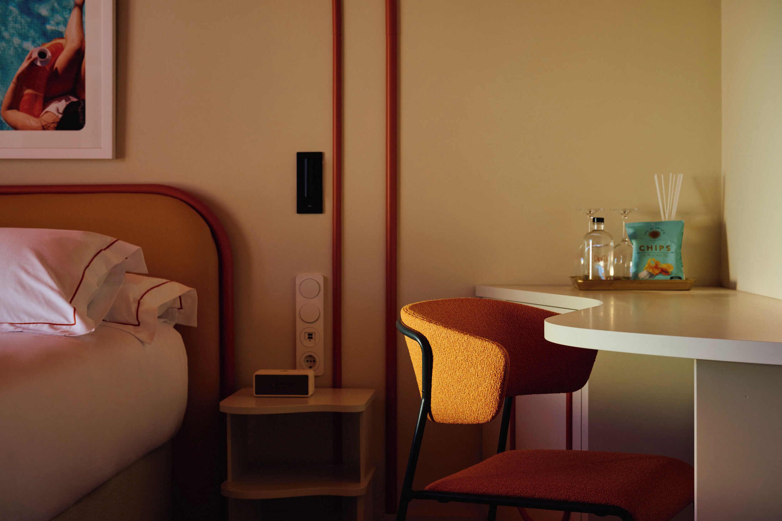 A hotel room with a bed, a desk, a chair, a nightstand, and a wall-mounted shelf with toiletries.
