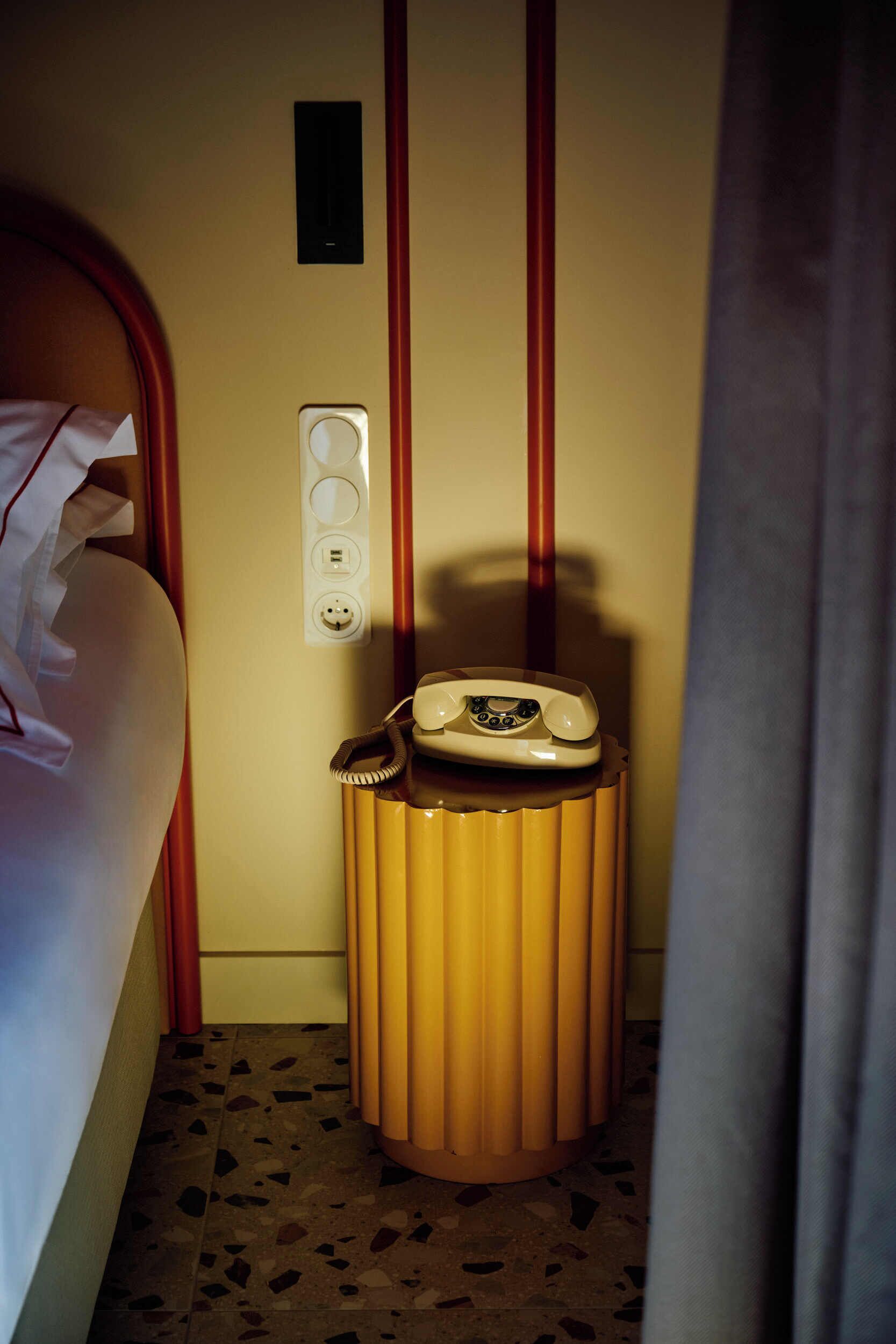 Habitación de hotel con cama, teléfono sobre una mesita, enchufe y un interruptor de luz en la pared.