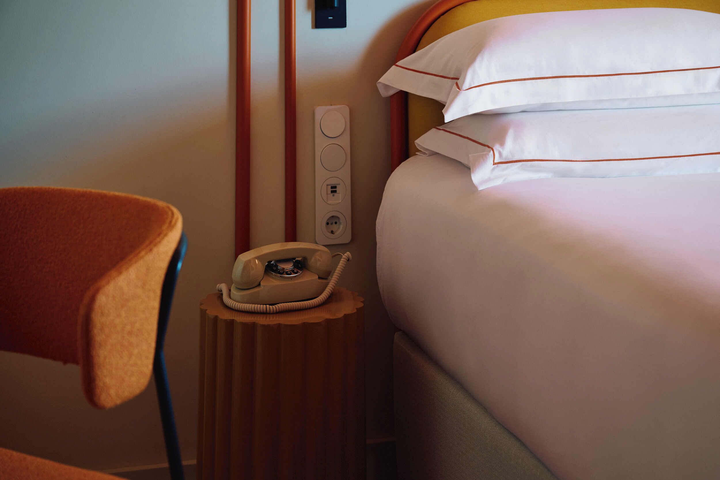Habitación de hotel con cama, una silla, una mesita de noche con teléfono y un enchufe de pared.
