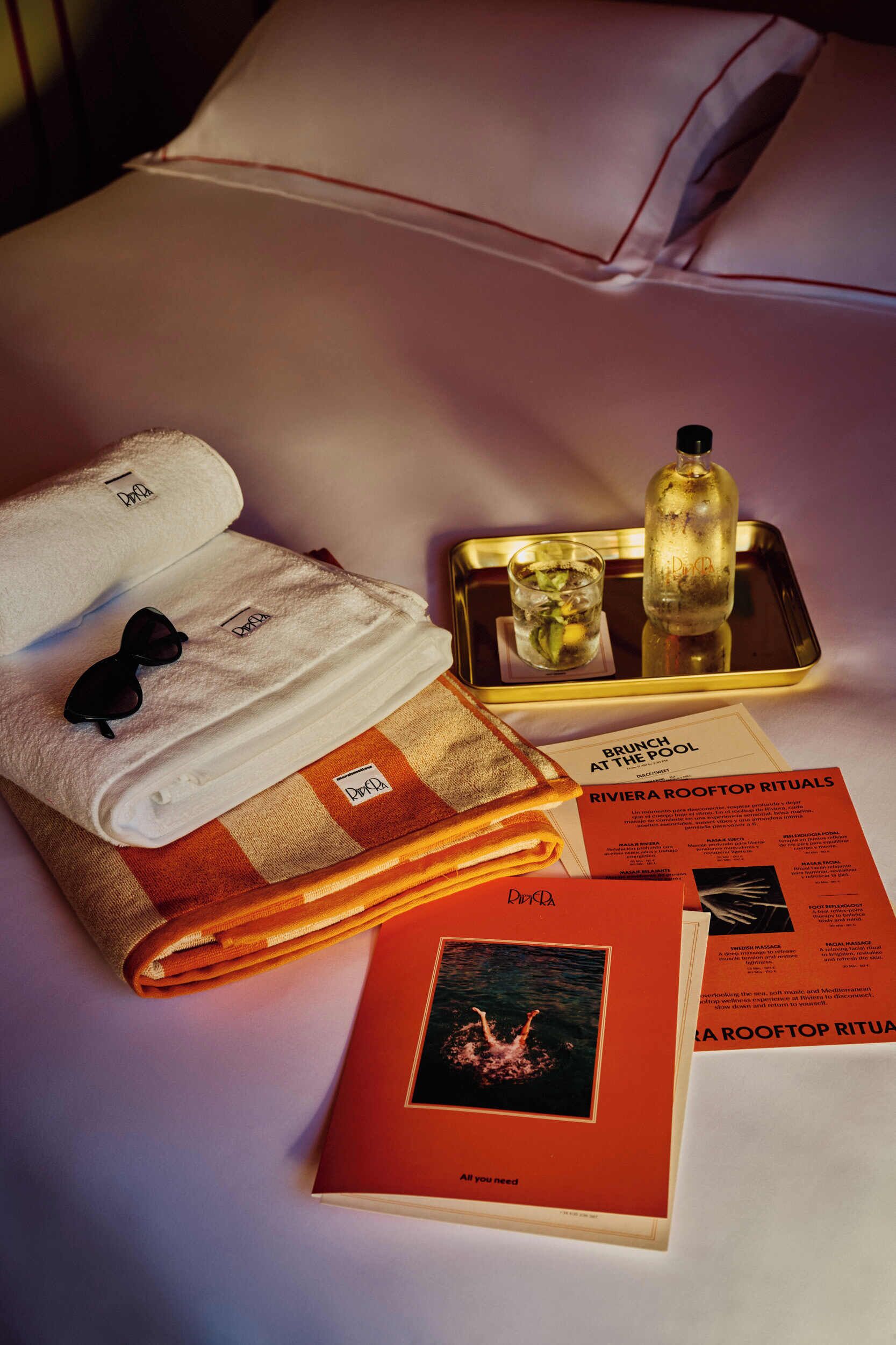 Une chambre d’hôtel bien rangée, avec un lit, un plateau sur lequel se trouvent un verre et une bouteille, une serviette pliée, des lunettes de soleil et des brochures posées sur le lit.