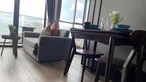 A modern living room with a sofa, a dining table, and a chair. There is a view of the city through large windows. - Sea View High Floor - Breathtaking Views (Pattaya)
