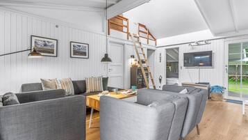 Moderni olohuone, jossa on parvelle sijoitettu makuuhuone, valkoiset seinäpaneelit, puinen tikapuu, televisio ja suuri ikkuna.