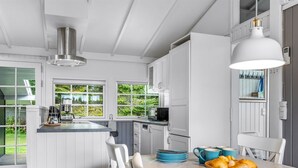 A modern kitchen with white cabinetry, a dining table with blue plates and a basket of bread, and a view of greenery outside.