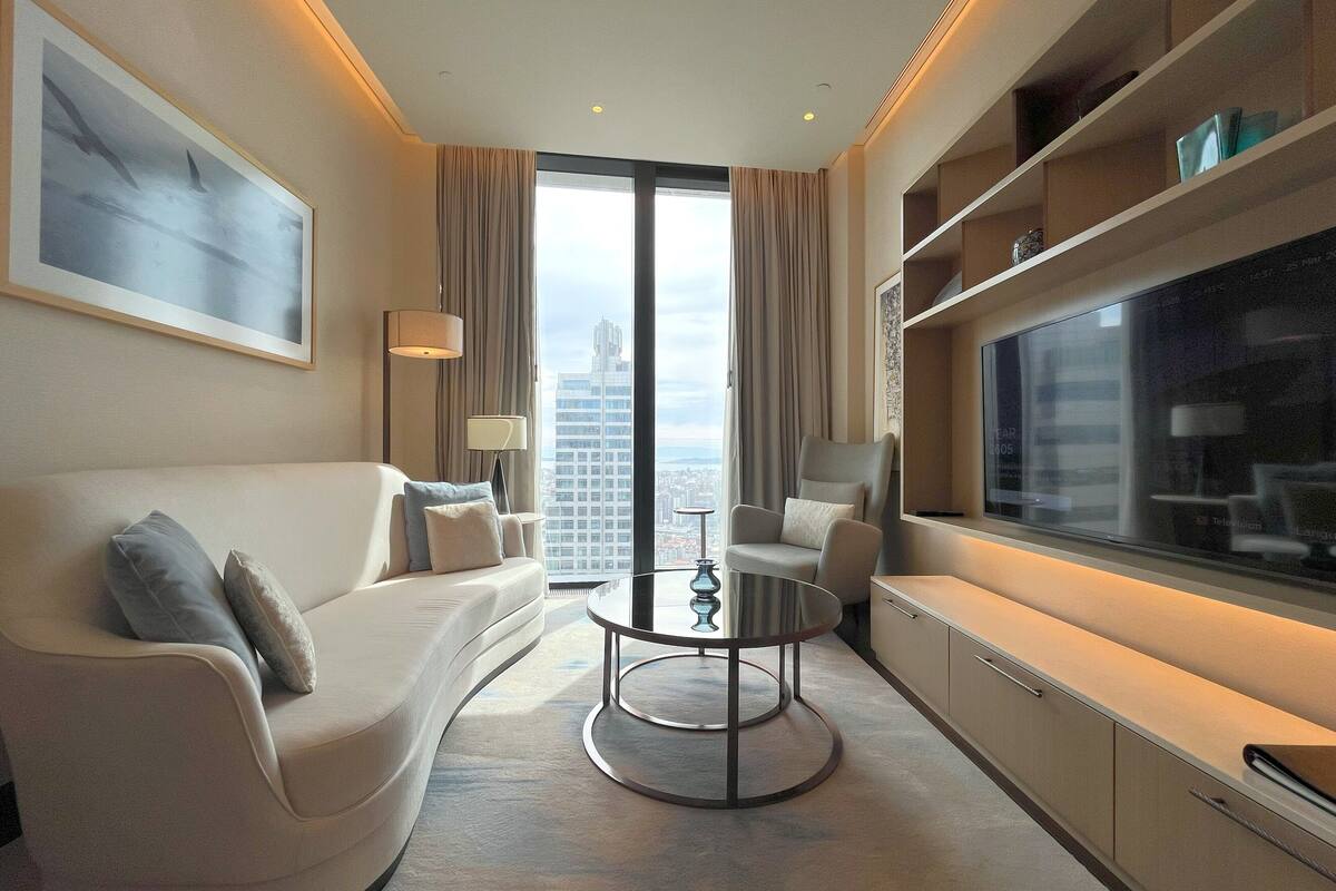 A modern hotel room with a curved white sofa, a round coffee table, a TV on a cabinet, and a large window with a city view.