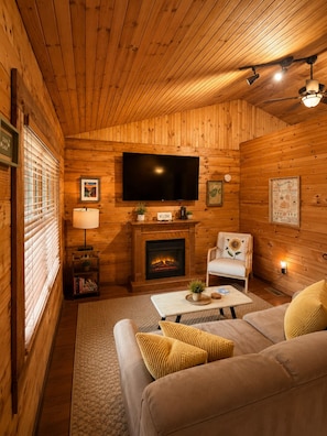 A cozy cabin interior with a fireplace, a television mounted on the wall, a sofa, and a small table.