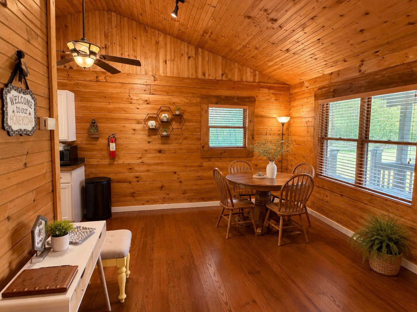 A wooden interior with a dining table, chairs, a ceiling fan, and a window with blinds.