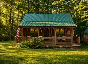 A wooden cabin with a green roof, a porch with seating, and an American flag.