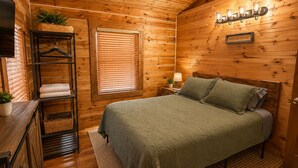 Un dormitorio de cabaña de madera acogedor con una cama, una mesita de noche, una lámpara, un estante con toallas y una ventana con persianas.