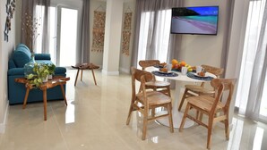 A modern living room with a blue sofa, wooden chairs, a dining table with fruit and a coffee cup, and a TV on the wall.