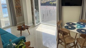 A modern living room with a large sliding glass door leading to a balcony, a dining area with wooden chairs and blue placemats, and a television mounted on the wall.