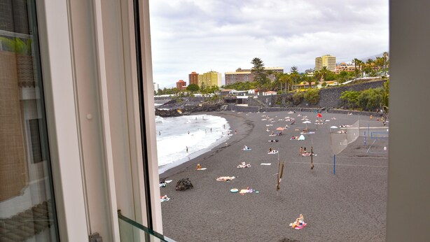 A beach with umbrellas and people, a rocky shoreline, and buildings in the background.