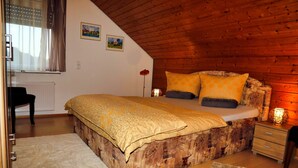 Un dormitorio acogedor con techo de madera, una cama con ropa de cama estampada, una mesita de noche con lámpara y un radiador.
