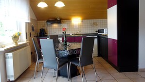 Una cocina con una isla central, dos sillas, una mesa, un refrigerador, un microondas y un ventanal con cortinas.