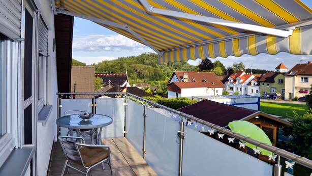 Un balcón con toldo a rayas, una mesa con sillas y vistas a casas y vegetación al fondo.