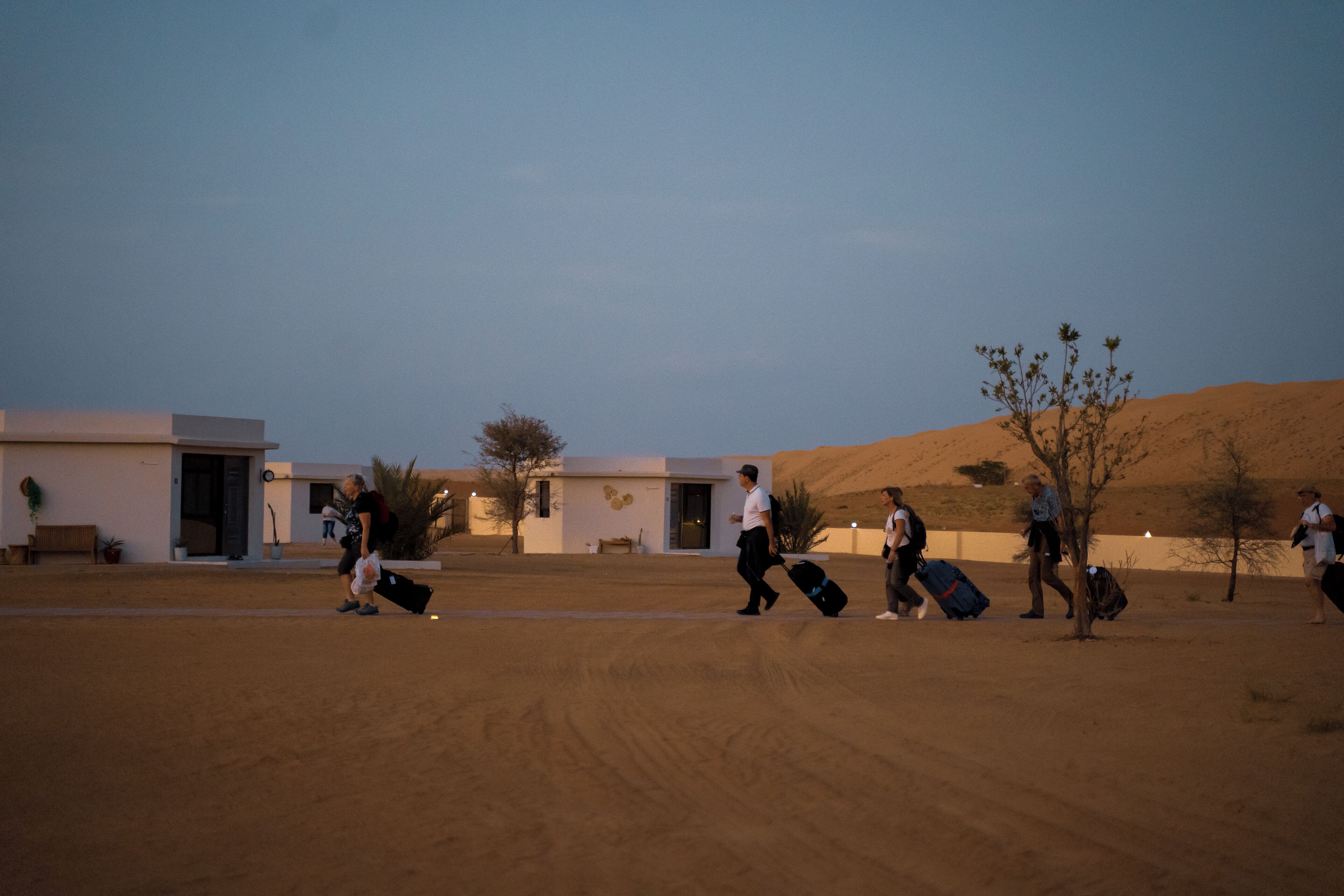 A desert landscape with a few buildings and people walking with luggage.