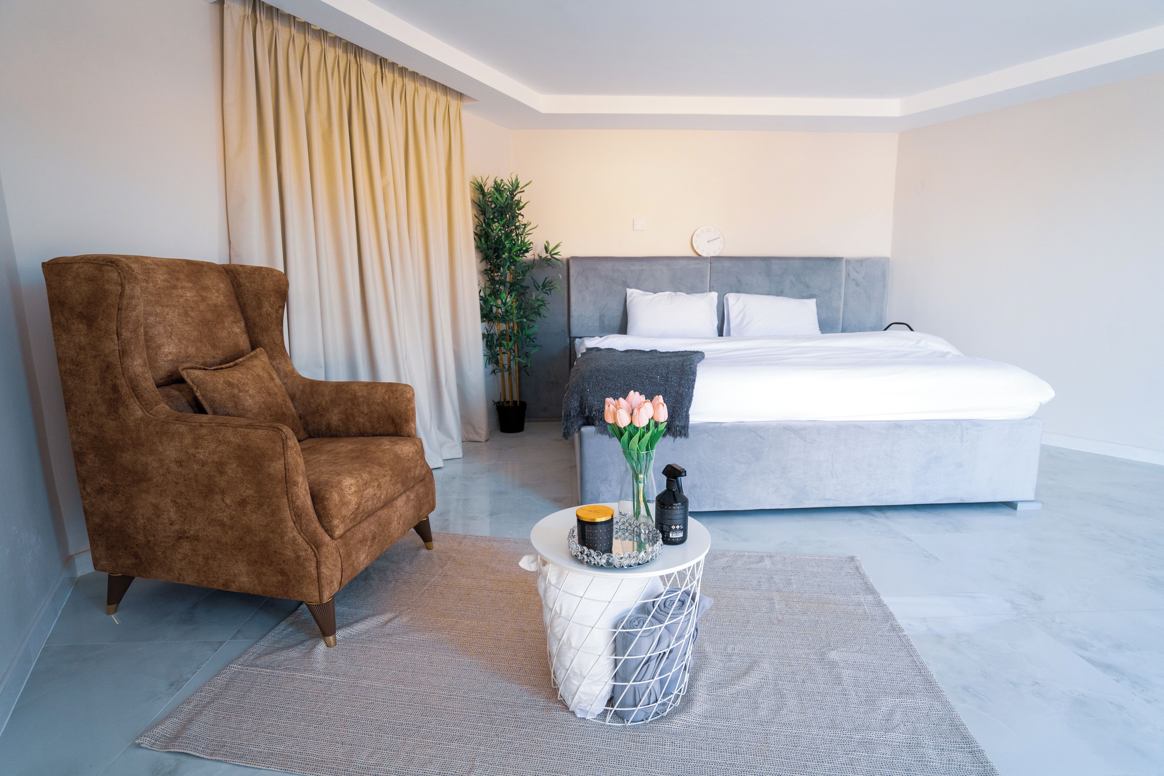 A modern hotel room with a large bed, a brown armchair, a small round table with a vase of flowers, and a potted plant.
