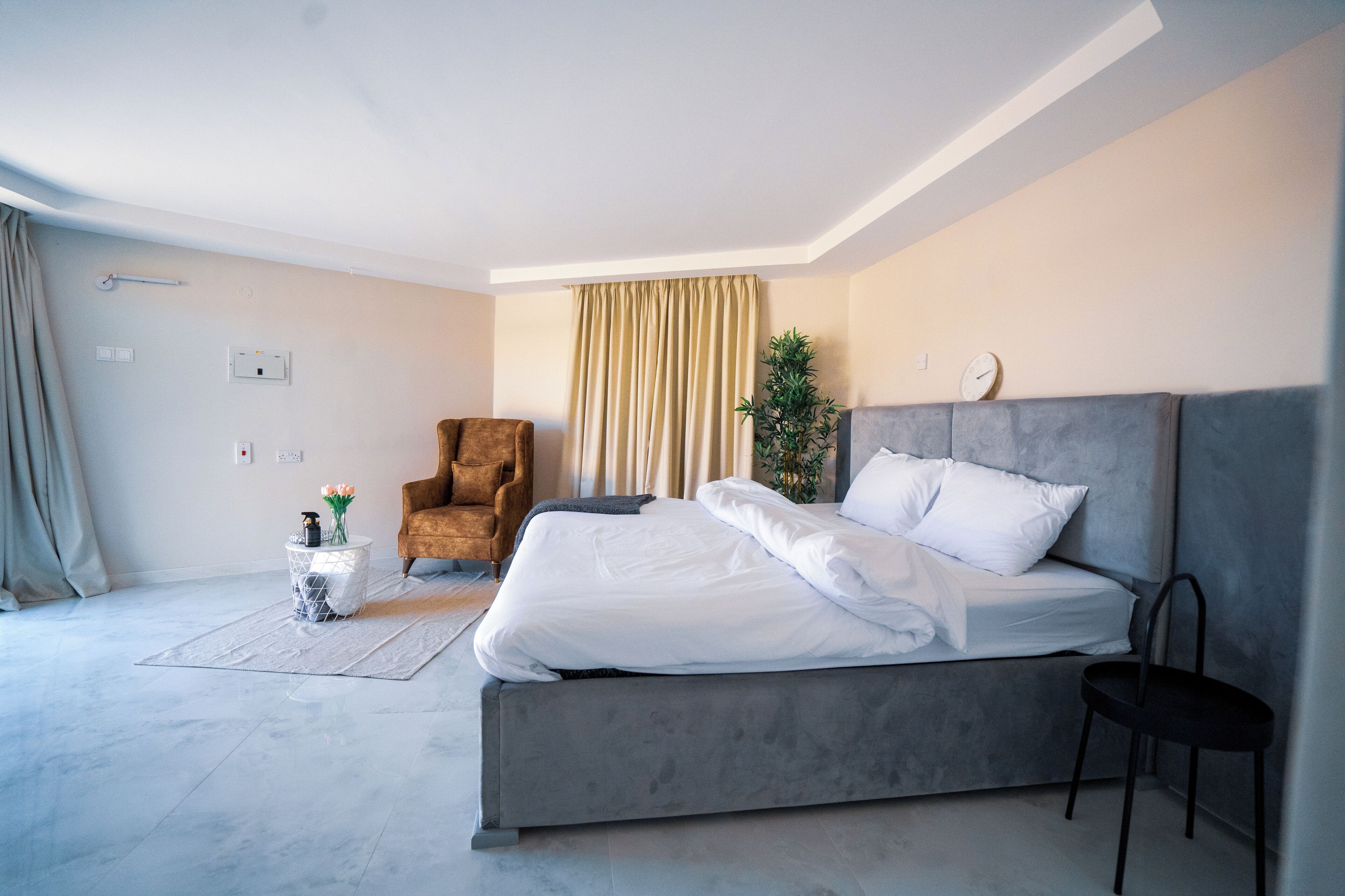 A modern bedroom with a large bed, a chair, a small table with flowers, and a window with curtains.